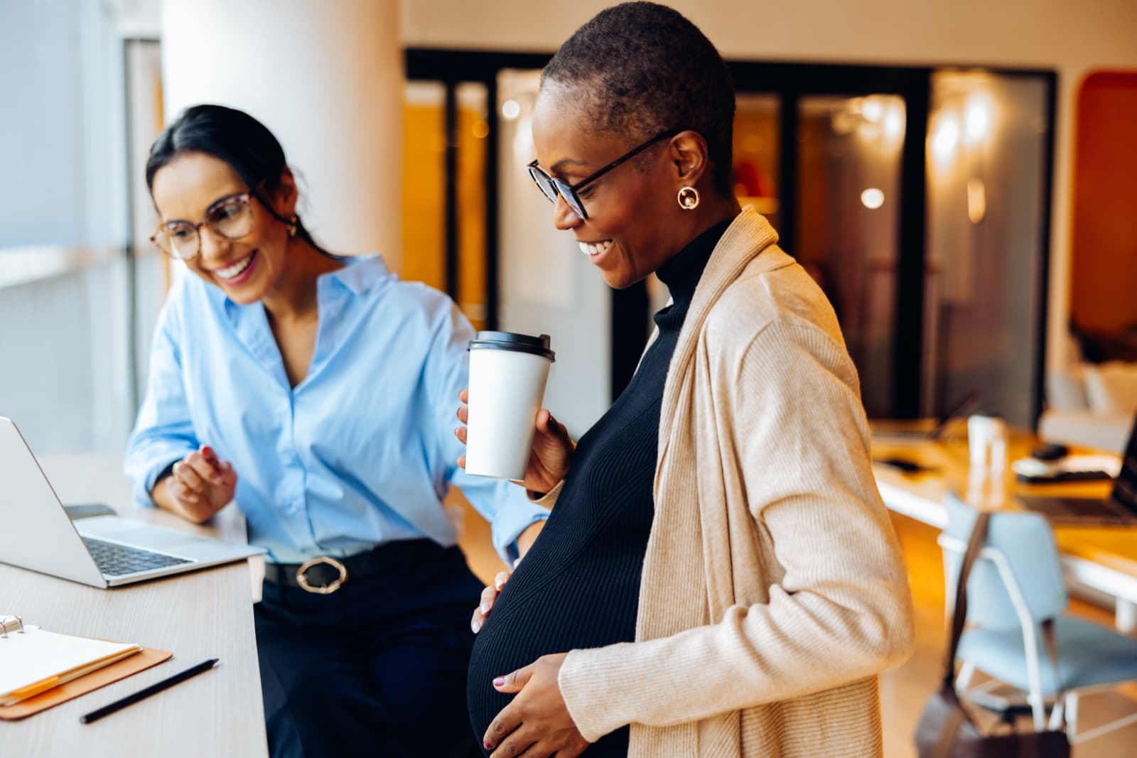 A smiling pregnant woman holding a coffee cup chats with a colleague at her laptop