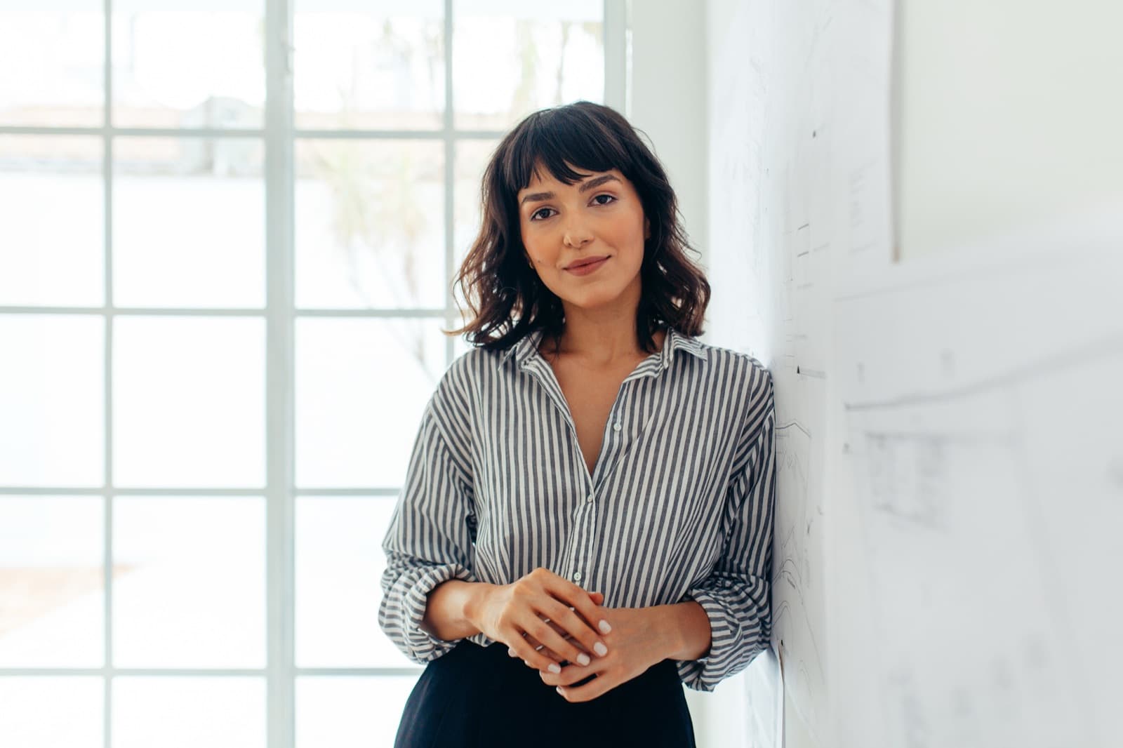 A smiling woman in a striped shirt leans against a wall in a bright office